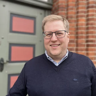 Ein Mann mit dunklem Pullover, Brille und blonden Haaren lächelt in die Kamera